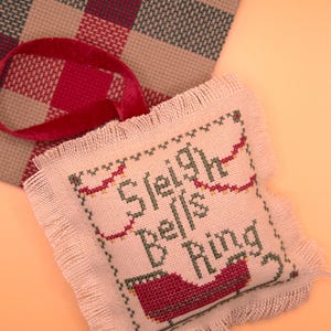 以下が含まれることがあります： フリンジの付いたピンクの布製のオーナメントで、"Sleigh Bells Ring"とクロスステッチで書かれています。オーナメントは赤と緑の糸で飾られており、赤いベルベットのリボンが付いています。