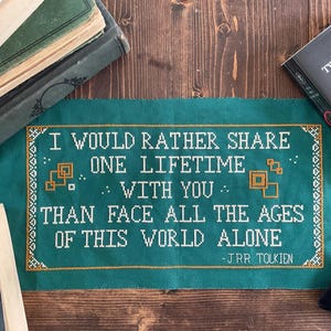 May include: A teal cross-stitch with the quote "I would rather share one lifetime with you than face all the ages of this world alone" by J.R.R. Tolkien. Books and a small black bag are in the background.