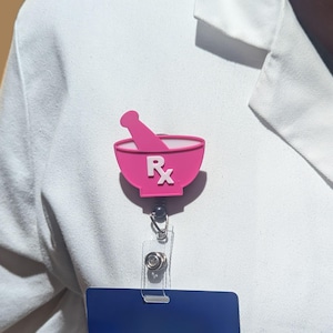 May include: A pink plastic badge holder with a mortar and pestle design and the letters "RX" in white. The badge holder is attached to a retractable badge reel with a blue and silver ID card.