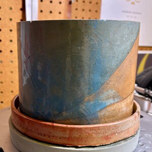 May include: A ceramic planter with a blue, brown, and tan glaze. The planter has a wide, round base and a slightly flared top.