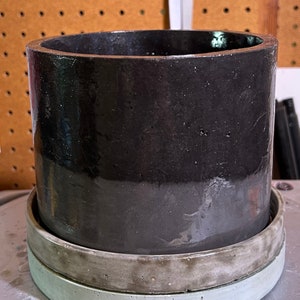 May include: A black ceramic planter with a gray concrete saucer. The planter has a smooth, glossy finish.