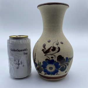 May include: A beige ceramic vase with a brown rim and a painted design of a bird and flowers in shades of blue, brown, and green. The vase is approximately 18 cm tall.
