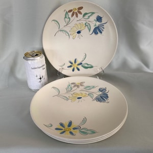 May include: Off-white ceramic plates with a floral design in yellow, blue, and brown. The plates have a simple, elegant design with a vintage aesthetic. A can of GoldenSparkle Shop soda is next to the plates.