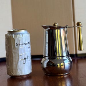 May include: A silver-coloured metal moka pot with a polished finish, featuring a gold-toned handle and a matching gold-toned knob on the lid. Beside it stands a cylindrical beverage can wrapped in a white label with the text GoldenSquareMile Shop.