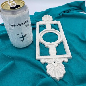 Peut inclure: Une décoration murale blanche ornée, avec un centre ovale, à côté d'une canette enveloppée dans une manchon en papier. Le manchon porte le texte "GoldenSquareMile Shop" et un motif d'éclat d'étoile. Le fond est en tissu turquoise.
