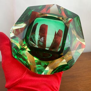 May include: A faceted, emerald green glass ashtray with a square interior and a circular base. The ashtray has a geometric design with red and gold accents. The object is held by a red gloved hand.