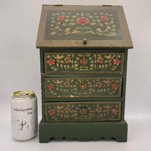 May include: A small, green wooden cabinet with three drawers and a hinged top. The cabinet is decorated with floral and bird designs in red, gold, and white. The cabinet has a decorative base and a small metal knob. A can is next to the cabinet.
