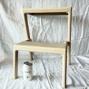 May include: A beige, modern chair with a rectangular seat and back. The chair has four straight legs and a minimalist design. A white can with black text and a graphic is next to the chair.