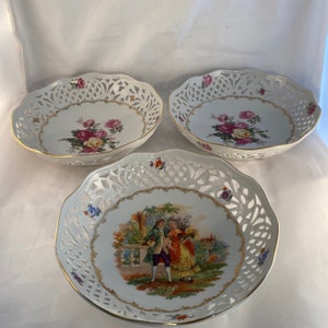May include: Three white porcelain bowls with intricate lace-like designs and gold trim. Two bowls feature floral designs, while the third bowl features a romantic scene of a couple in a garden.