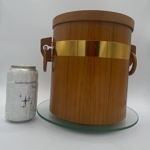 Peut inclure: Seau à glace vintage en bois avec une bande dorée et des poignées en bois. Le seau cylindrique repose sur une assiette en verre transparente. Une canette avec le texte "GoldenSquareMile Shop" est à côté du seau.
