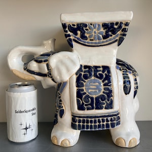 May include: A white ceramic elephant garden stool featuring deep blue and light blue floral and geometric patterns. The elephant has a flat top, a trunk curled upward, and is positioned next to a silver aluminium can with a white label that reads GoldenSquareMile Shop with four-pointed star icons.