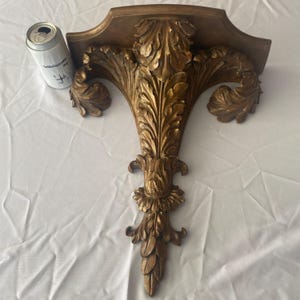Peut inclure: Étagère murale dorée ornée avec un design symétrique détaillé. L'étagère présente un motif floral central vertical avec des feuilles enroulées et une base décorative. L'étagère est faite d'un matériau solide et a un dessus incurvé.