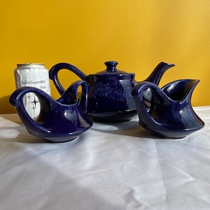 Peut inclure: Un service à thé bleu cobalt vintage, comprenant une théière avec couvercle et deux crémier. L'ensemble a une finition émaillée mouchetée. Une canette avec le texte "GoldenSquareNile Shop" est visible en arrière-plan.