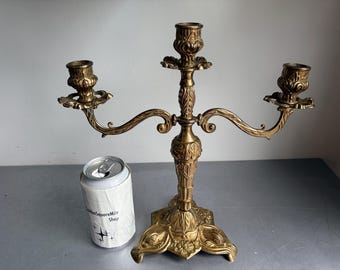 May include: A gold-toned metal candelabra with three candle holders, featuring ornate embossed detailing on the central stem and base. A silver-colored aluminum beverage can with a white label reading GoldenSquareMile Shop sits beside it.