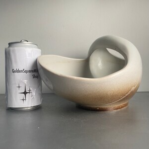 May include: A white ceramic bowl with a brown glaze on the bottom. The bowl has a unique shape with a handle on one side. The bowl is ideal for serving snacks or fruit.
