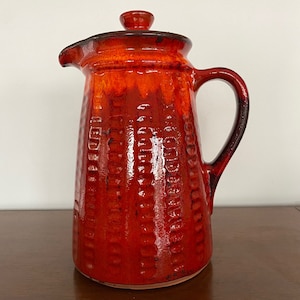 May include: A red ceramic jug with a lid and a textured surface. The jug has a wide base and a narrow spout.