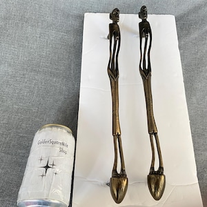 May include: Two bronze-coloured serving utensils with human figure handles. The utensils are on a white surface, with a can of Golden Square Mile Shop in the lower left corner. The utensils have a unique, artistic design.