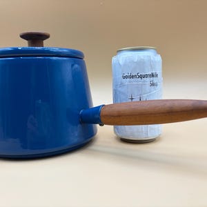 May include: A blue enamel saucepan with a wooden handle and lid. The saucepan has a wooden knob on the lid. A can with the text "GoldenSquareMile Shop" is next to the saucepan.