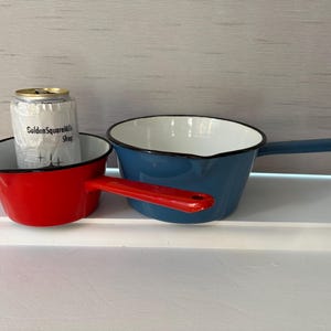 Peut inclure: Deux casseroles en émail, une rouge et une bleue, avec des intérieurs blancs et des bords noirs. La casserole rouge a une poignée rouge, et la casserole bleue a une poignée bleue. Une canette enveloppée de papier se trouve à l'intérieur de la casserole rouge.