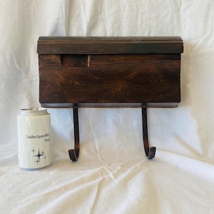 May include: A dark brown wooden wall-mounted mail holder with a curved top and two metal hooks. A white can with the text "GoldenSquareMill Shop" and a star design is on the left.