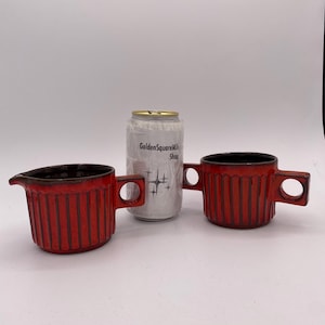 May include: Two red ceramic mugs with black interiors and handles. The mugs have vertical ribbed detailing. A white can with text is between the mugs. The mugs are likely for coffee or tea.