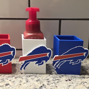 May include: A red, white, and blue soap dispenser and holders. The holders are red, white, and blue, each with a Buffalo Bills logo. The soap dispenser is red and sits in a white holder. The items are on a speckled countertop.