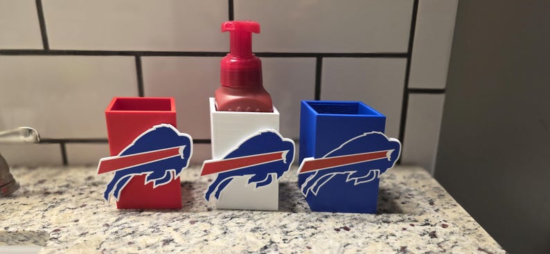 May include: A red, white, and blue soap dispenser and holders. The holders are red, white, and blue, each with a Buffalo Bills logo. The soap dispenser is red and sits in a white holder. The items are on a speckled countertop.