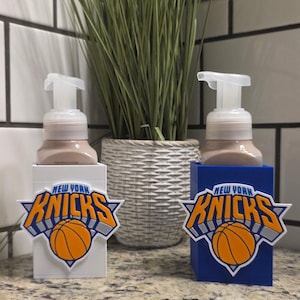 May include: Two rectangular soap dispensers with pump tops, one white and one blue. Each features the New York Knicks basketball team logo on the front, displaying the text New York Knicks in blue and orange with an orange basketball graphic.
