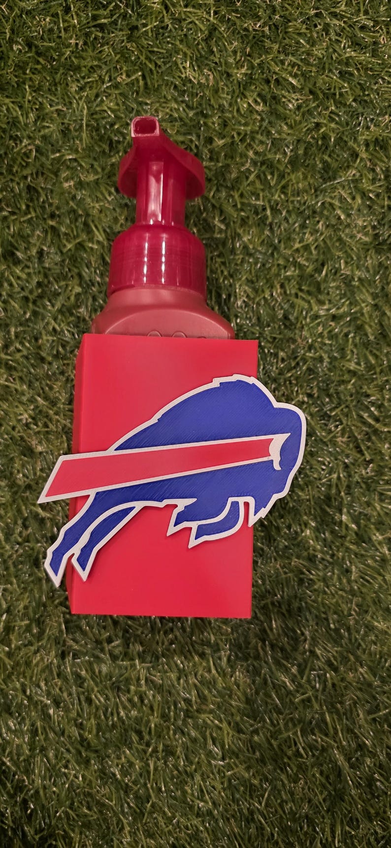 May include: A red soap dispenser with a red pump and a blue and red Buffalo Bills logo. The dispenser is on a green surface.