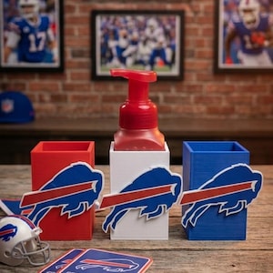 May include: A set of three rectangular containers in red, white, and blue, each featuring a Buffalo Bills logo. A red soap pump sits in the white container. A miniature football helmet and two square coasters with the team logo are included.