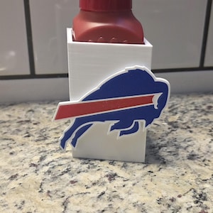 May include: A red soap dispenser with a white holder featuring the Buffalo Bills logo. The logo is a blue buffalo with a red stripe. The dispenser is on a speckled countertop.