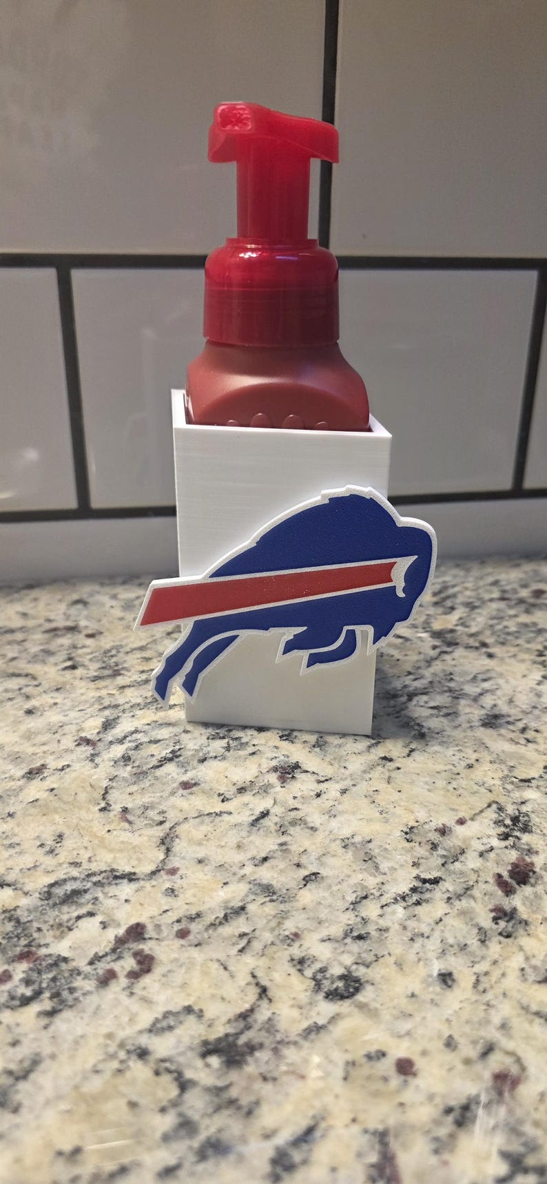 May include: A red soap dispenser with a white holder featuring the Buffalo Bills logo. The logo is a blue buffalo with a red stripe. The dispenser is on a speckled countertop.