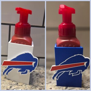 May include: A red soap dispenser with a red pump sits in a white holder with a Buffalo Bills logo. The logo features a blue buffalo with a red stripe. The holder is on a countertop.