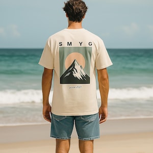 Op de afbeelding: Beige t-shirt met een bergafbeelding en de tekst "SMYG since 2021". Het t-shirt wordt gecombineerd met een spijkerbroek. De persoon staat op een zandstrand met de oceaan op de achtergrond.