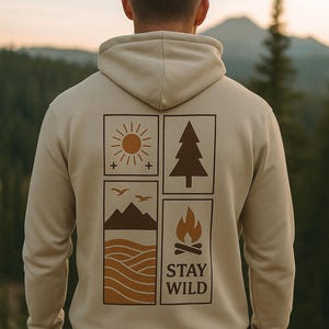 Könnte beinhalten: Beigefarbener Kapuzenpullover mit einem Naturmotiv auf dem Rücken. Das Design zeigt eine Sonne, einen Baum, Berge und ein Lagerfeuer mit dem Schriftzug "STAY WILD". Der Hoodie hat eine Kapuze und eine lockere Passform.