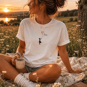 Könnte beinhalten: Ein weißes kurzärmeliges T-Shirt mit einem kleinen Grafikdruck, der die Silhouette eines Kindes zeigt, das nach einem herzförmigen Luftballon greift, daneben das Wort love in schwarzer Schreibschrift. Das Shirt hat einen Rundhalsausschnitt.