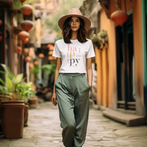 Könnte beinhalten: Eine Frau trägt ein weißes T-Shirt mit dem Wort "happy" vertikal in bunten Buchstaben gedruckt. Das Shirt hat die Wörter "care", "hope", "love" und "smile" vertikal neben dem Wort "happy" gedruckt. Sie trägt einen breitkrempigen Hut und eine grüne Hose. Sie geht eine Straße entlang, die mit orangefarbenen Laternen gesäumt ist.