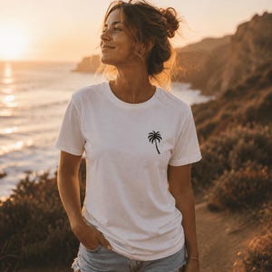 May include: White t-shirt with a black palm tree graphic. The short-sleeved shirt is worn with denim shorts. The image is taken outdoors with a sunset in the background. The person is standing on a path near the ocean.