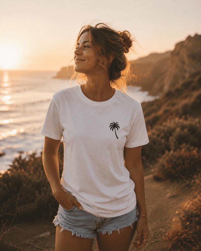 May include: White t-shirt with a black palm tree graphic. The short-sleeved shirt is worn with denim shorts. The image is taken outdoors with a sunset in the background. The person is standing on a path near the ocean.