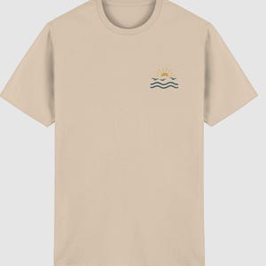 Pu&ograve; includere: Una t-shirt beige a maniche corte con una piccola grafica sulla parte superiore sinistra del petto. La grafica raffigura un sole giallo che sorge sopra onde marine blu scuro con tre piccoli uccelli blu scuro che volano tra il sole e le onde.