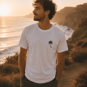 May include: White t-shirt with a black palm tree graphic. The short-sleeved shirt is made of a soft material. The person is standing outside near the ocean at sunset, wearing dark green shorts.