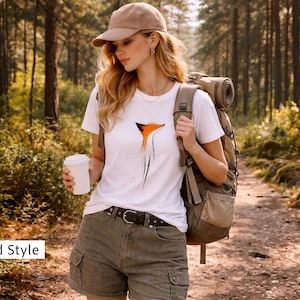 May include: A white t-shirt featuring a stylised orange and black fox graphic, paired with olive green cargo shorts, a brown leather belt, a tan baseball cap, and a large hiking backpack. The shirt is worn with a white cup in one hand.