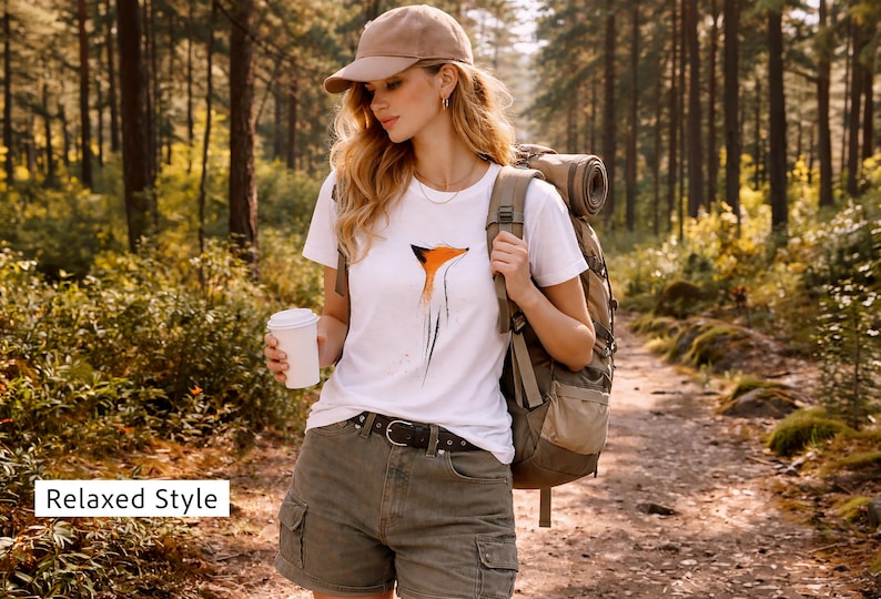 May include: A white t-shirt featuring a stylised orange and black fox graphic, paired with olive green cargo shorts, a brown leather belt, a tan baseball cap, and a large hiking backpack. The shirt is worn with a white cup in one hand.