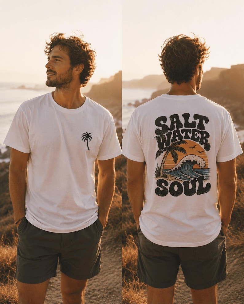 May include: White t-shirt with a palm tree graphic on the front and "Salt Water Soul" design on the back. The back design features a palm tree, waves, and a sunset. The person is wearing dark green shorts.