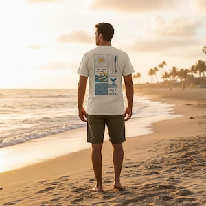 Pu&ograve; includere: Una t-shirt a maniche corte color crema con una grafica rettangolare sul retro. Il disegno include un sole, una tavola da surf, uccelli, onde dell'oceano e una coda di balena, con il testo "LIFE IS GOOD" stampato in blu scuro.