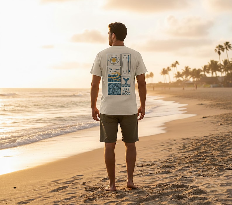 Pu&ograve; includere: Una t-shirt a maniche corte color crema con una grafica rettangolare sul retro. Il disegno include un sole, una tavola da surf, uccelli, onde dell'oceano e una coda di balena, con il testo "LIFE IS GOOD" stampato in blu scuro.