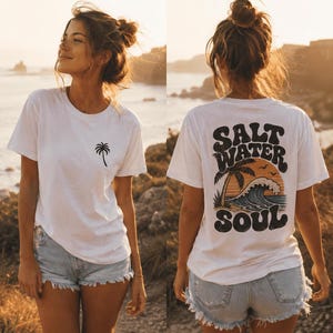 May include: White t-shirt with a palm tree graphic on the front and "SALT WATER SOUL" design on the back. The shirt is paired with denim shorts. The image is taken outdoors with a scenic background.