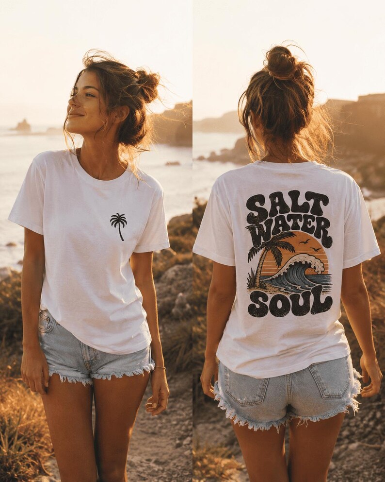 May include: White t-shirt with a palm tree graphic on the front and "SALT WATER SOUL" design on the back. The shirt is paired with denim shorts. The image is taken outdoors with a scenic background.