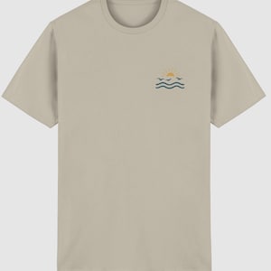 Pu&ograve; includere: Una t-shirt beige a maniche corte con una piccola grafica sul petto sinistro. La grafica raffigura un sole giallo che sorge sopra tre linee ondulate color verde acqua scuro che rappresentano l'acqua, con tre piccoli uccelli.