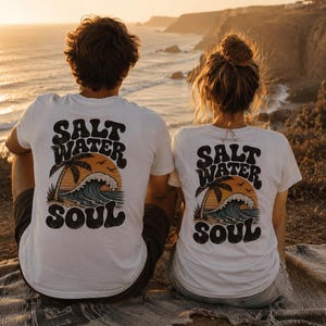 May include: Two white t-shirts with the text "SALT WATER SOUL" on the back, featuring a palm tree, waves, and a sunset design. The shirts are worn by two people sitting on a blanket, overlooking the ocean at sunset.